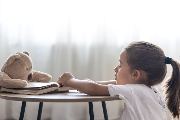 Маленькая девочка играет со своим плюшевым мишкой и книгой, учит его читать, играет в школе.