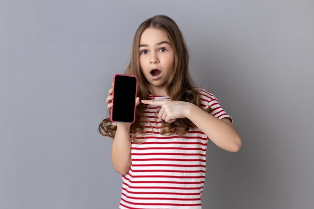 Little girl pointing finger at smartphone with white empty display shocked by application