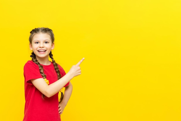 A little girl points her finger at an empty space for your advertisement on a yellow isolated background A beautiful child is smiling in a red Tshirt with two pigtails Copy space