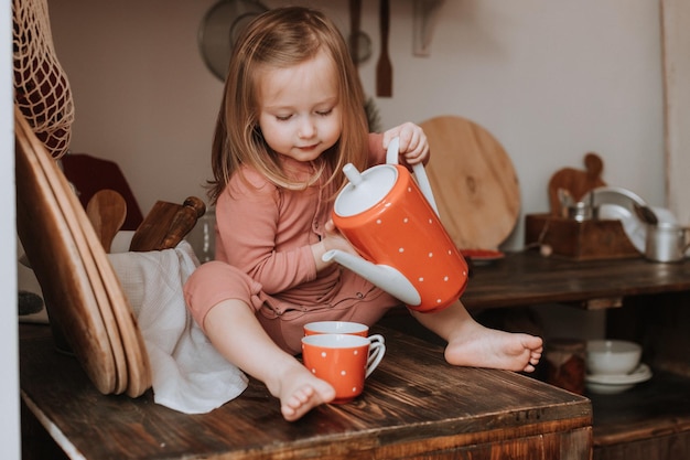 小さな女の子は、白いエンドウ豆の木製キッチンのティーポットの赤いセラミック皿からマグカップにお茶を注ぐ
