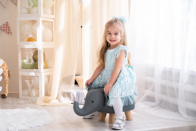 A little girl in princess dress sits on a wooden elephant toy at playroom