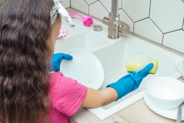 Little girl in protective gloves washing up white plate with foamy dishwashing sponge and dish soap in kitchen sink