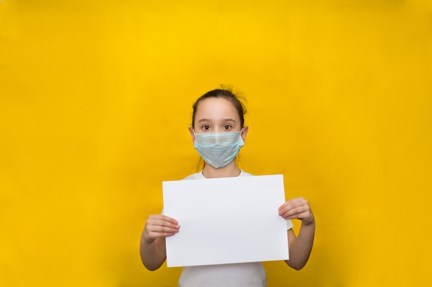 La bambina in una maschera protettiva tiene un foglio bianco su una parete gialla. Protezione contro il coronavirus