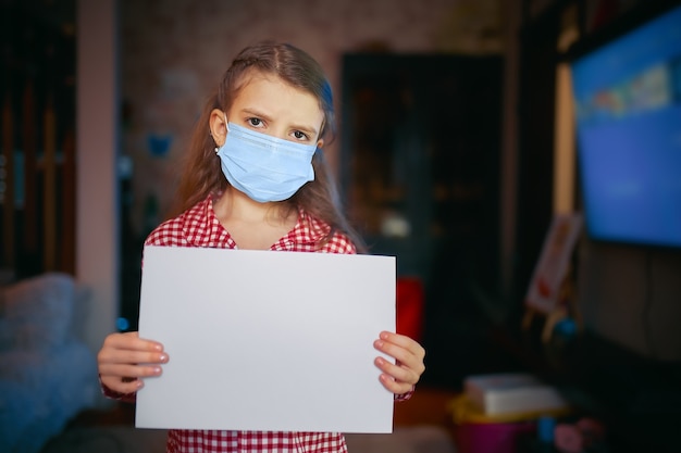 Little girl in a protective mask, pajamas holds a blank sheet of paper while standing in room at home, Protection against coronavirus