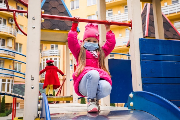 The little girl in a protective mask on the playground in the\
fall she sits on top of the slide and holds the crossbar