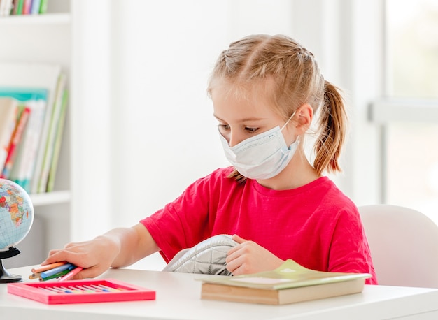 Little girl in protective mask at school lesson in bright classroom