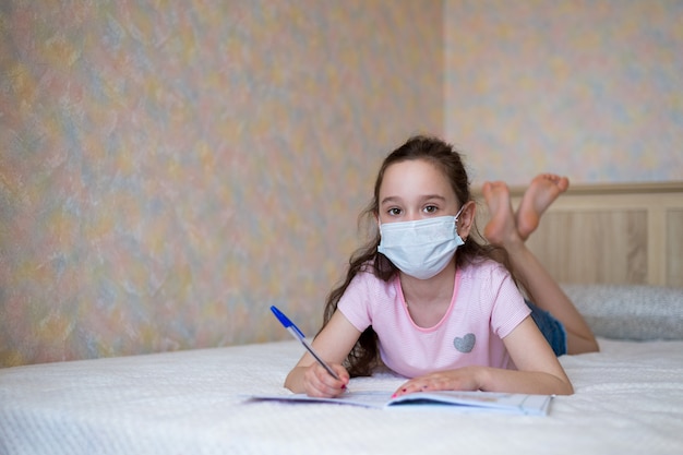 Photo little girl in a protective mask writes in a notebook during quarantine.