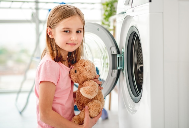 Маленькая девочка кладет плюшевую игрушку в открытую стиральную машину и улыбается
