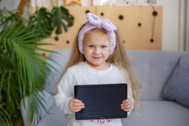 Little girl in pyjamas with a bandage on the eye at home on the sofa playing tablet