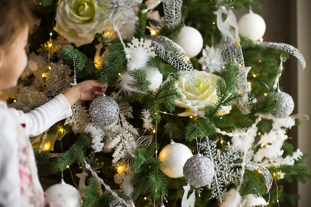 Una bambina raggiunge con la mano un giocattolo per l'albero di natale. decorazioni natalizie, in attesa di una vacanza e di un miracolo. nuovo anno. decorazioni bianche, rosa e blu, ghirlande di luci dorate