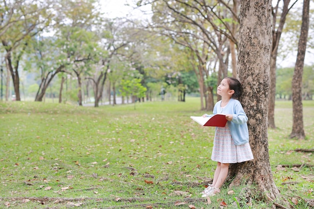 Little girl reading book in park lean against tree with looking up.