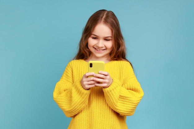 Bambina che legge il messaggio sullo smartphone e sorridente, soddisfatta dell'applicazione mobile per bambini, indossando un maglione giallo in stile casual. studio indoor girato isolato su sfondo blu.