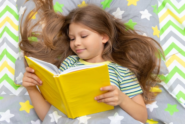 A little girl reads a book with interest in bed before going to bed.