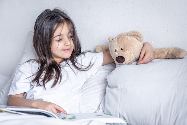 小さな女の子は朝、ベッドでテディベアと一緒に本を読みます