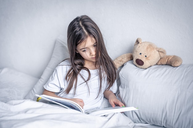 Little girl reads a book with a teddy bear in bed in the morning