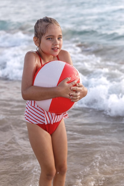 A little girl in a red bathing suit with a big inflatable ball in her hands is shivering from the cold after swimming in the sea