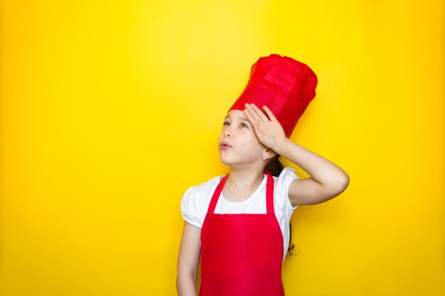 La bambina nella tuta di un cuoco rosso è stanca di cucinare, si asciuga la fronte con la mano sul giallo