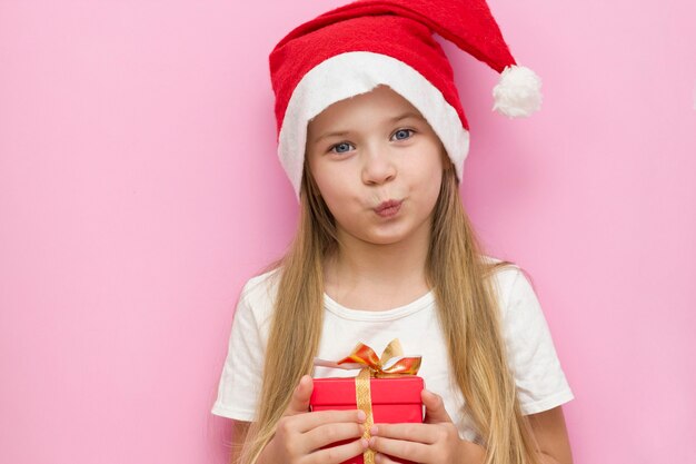 little girl in a red Christmas hat presents a red box with a gold bow, a gift. Smiles. The new year is 2021. Close up