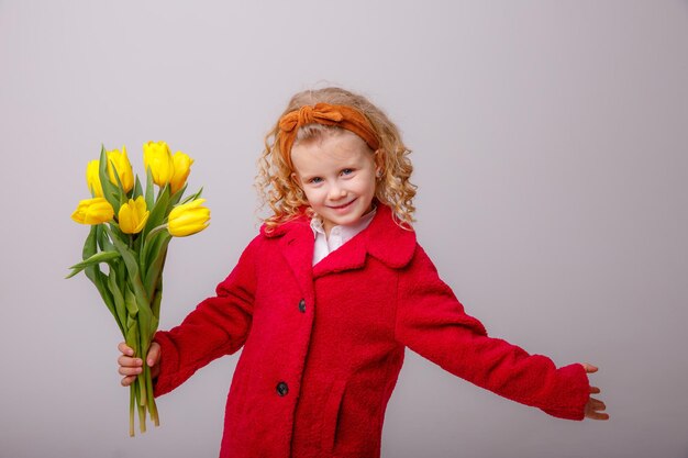 Una bambina con un cappotto rosso tiene un mazzo di fiori di tulipano primaverile su uno sfondo bianco