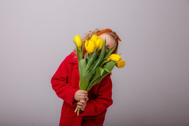 Una bambina con un cappotto rosso tiene un mazzo di fiori di tulipano primaverile su uno sfondo bianco