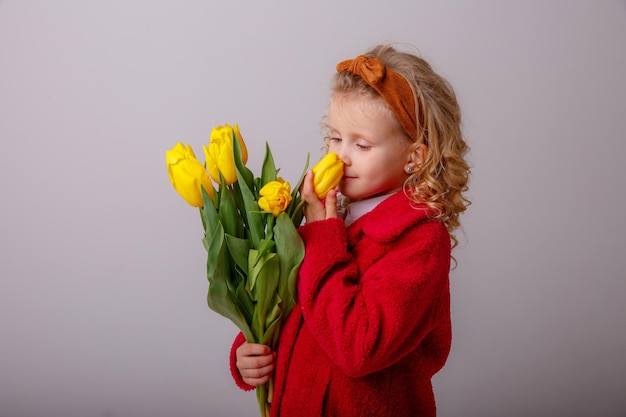 Una bambina con un cappotto rosso tiene un mazzo di fiori di tulipano primaverile su uno sfondo bianco