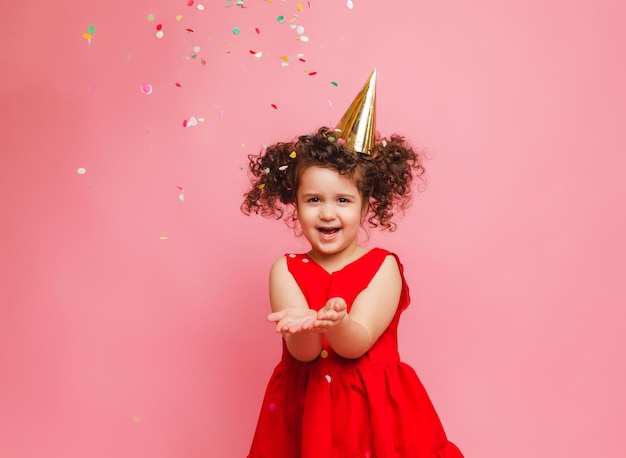 A little girl in a red dress celebrates her birthday blowing and catching confetti on a pink background