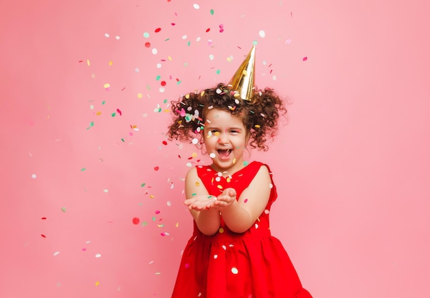 A little girl in a red dress celebrates her birthday blowing and catching confetti on a pink background