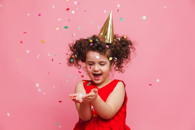 Photo a little girl in a red dress celebrates her birthday blowing and catching confetti on a pink background