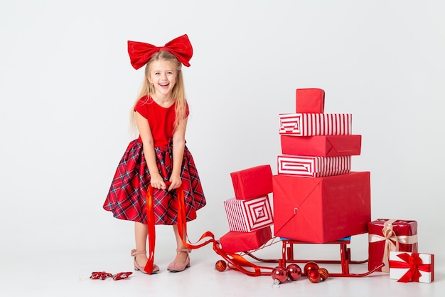 La bambina in un vestito rosso rotola una slitta con i regali per il nuovo anno. sfondo bianco, spazio per il testo. il concetto di natale