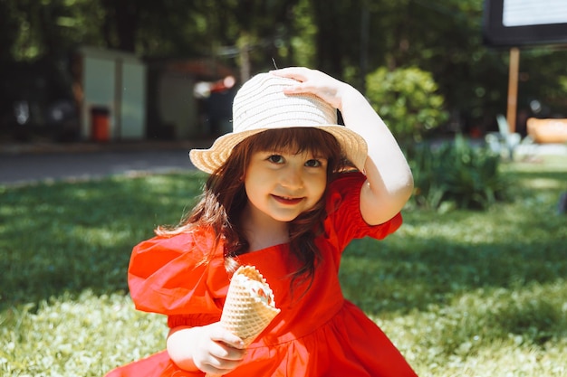 A little girl in a red dress and a straw hat eats ice cream in a cone in the park