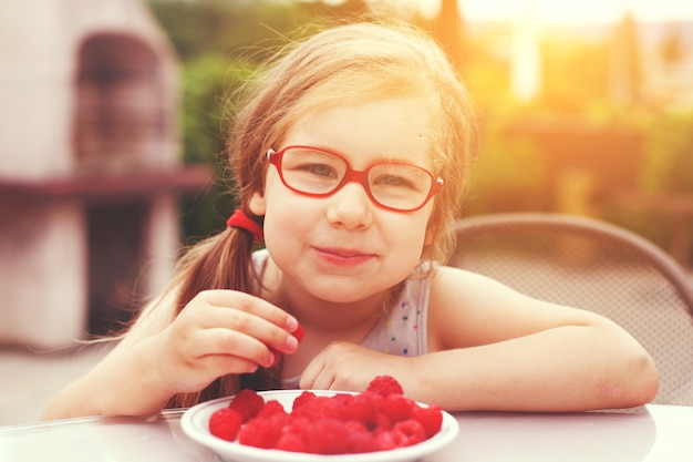 Foto bambina con gli occhiali rossi che mangia lamponi maturi in un caffè all'aperto