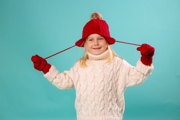 маленькая девочка в красной вязаной шапке, варежках и белом свитере улыбается на синей стене