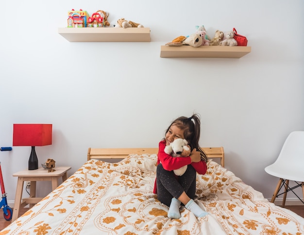 Little girl in red long neck t-shirt sweater hugging a teddy bear with red scarf.