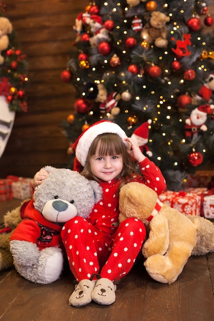 Little girl in red pajamas sitting with gifts by Christmas tree