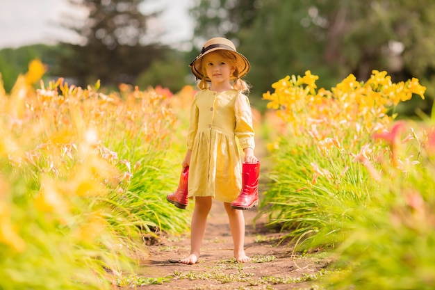 маленькая девочка в красных резиновых сапогах и соломенной шляпе поливает красные цветы в саду