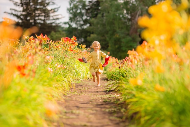 Bambina in stivali di gomma rossi e un cappello di paglia che innaffia i fiori d'innaffiatura rossi nel giardino