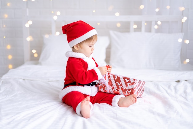 A little girl in a red Santa costume is sitting on a bed looking at New Years gifts in a red box