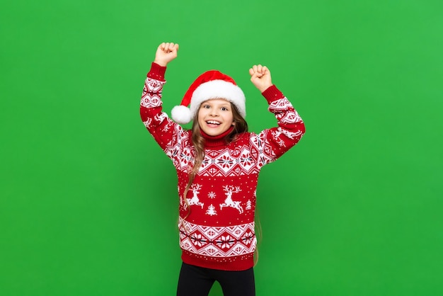 A little girl in a red sweater with reindeer and a Santa Claus hat waiting for the holiday on a green isolated background The concept of Christmas