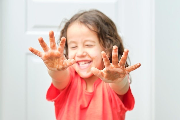 Bambina con una maglietta rossa sorridente con gli occhi chiusi con il viso sfocato e le mani tese in primo piano imbrattate di cioccolato