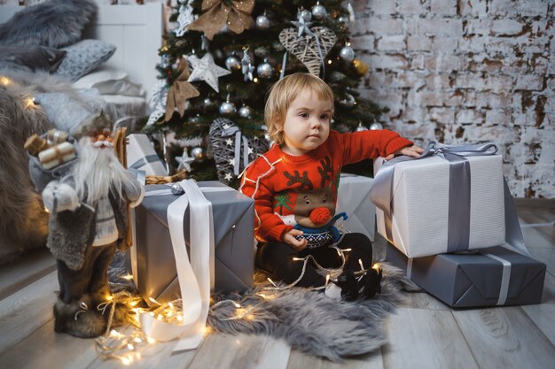 赤い暖かいセーターを着た少女が、おもちゃやプレゼントを持ってクリスマスツリーの下に座っています。幸せな子供時代。年末年始の雰囲気
