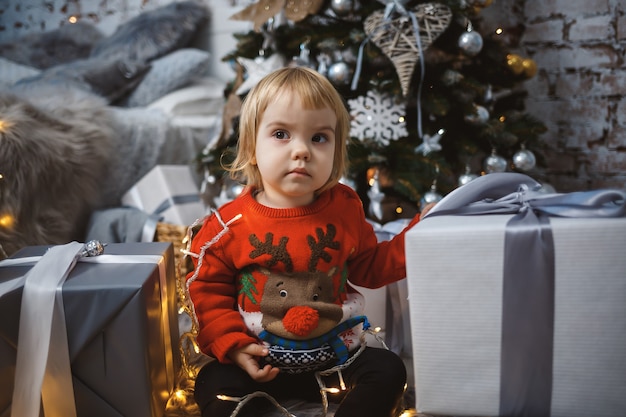 A little girl in a red warm sweater sits under a Christmas tree with toys and gifts. Happy childhood. New Year holiday atmosphere