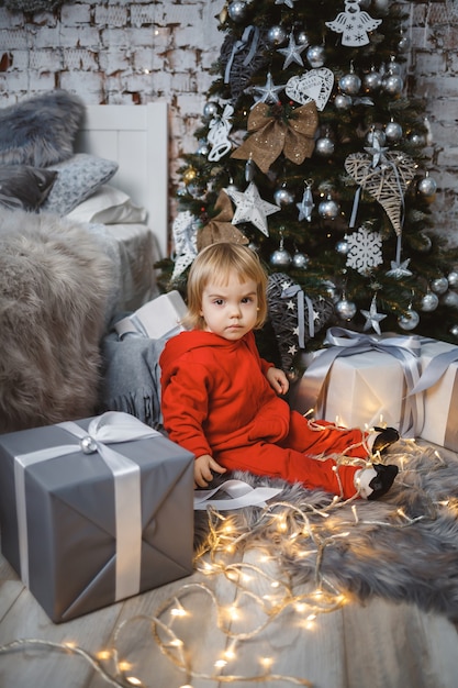 A little girl in a red warm sweater sits under a Christmas tree with toys and gifts. Happy childhood. New Year holiday atmosphere