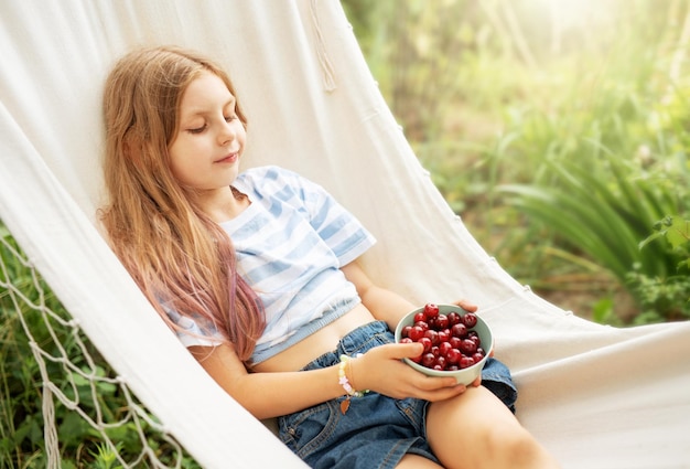 A little girl rests in a hammock and eats cherries in the summer