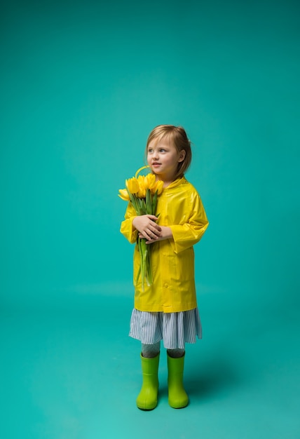 A little girl in rubber boots and a raincoat stands with yellow tulips on turquoise