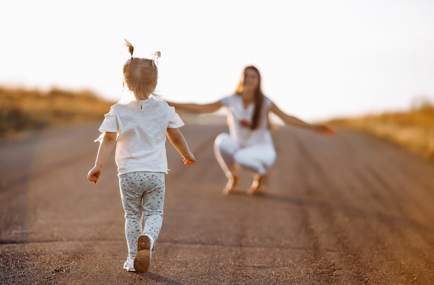 Little girl runs along the road to meet mom in the summer at sunset