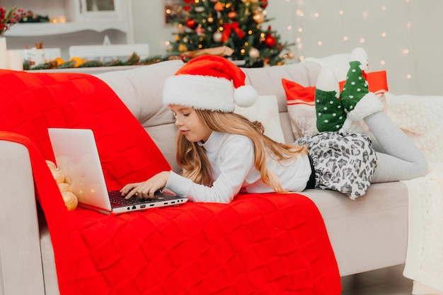 A little girl in a Santa Claus hat communicates by video call via a laptop, lying on the couch in the living room.