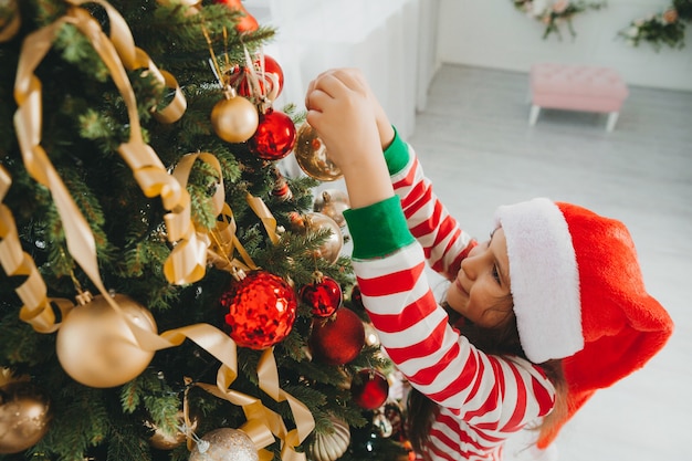 Little girl in santa claus hat decorates the tree with toys. A cute baby is preparing to celebrate Christmas.