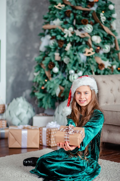 벽난로 옆에 앉아 크리스마스 트리 아래 선물을 가진 어린 소녀 산타 클로스 모자 선물을 풉니 다.