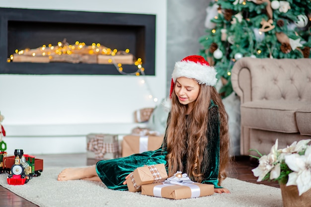 Little girl Santa Claus hat with gifts under Christmas tree sitting by the fireplace, unpacks gifts.