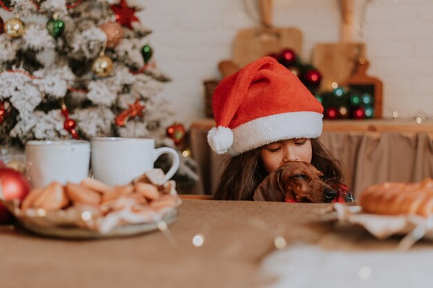 Little girl in a Santa hat and dwarf dachshund want to eat a plate of pastries and a Christmas cake